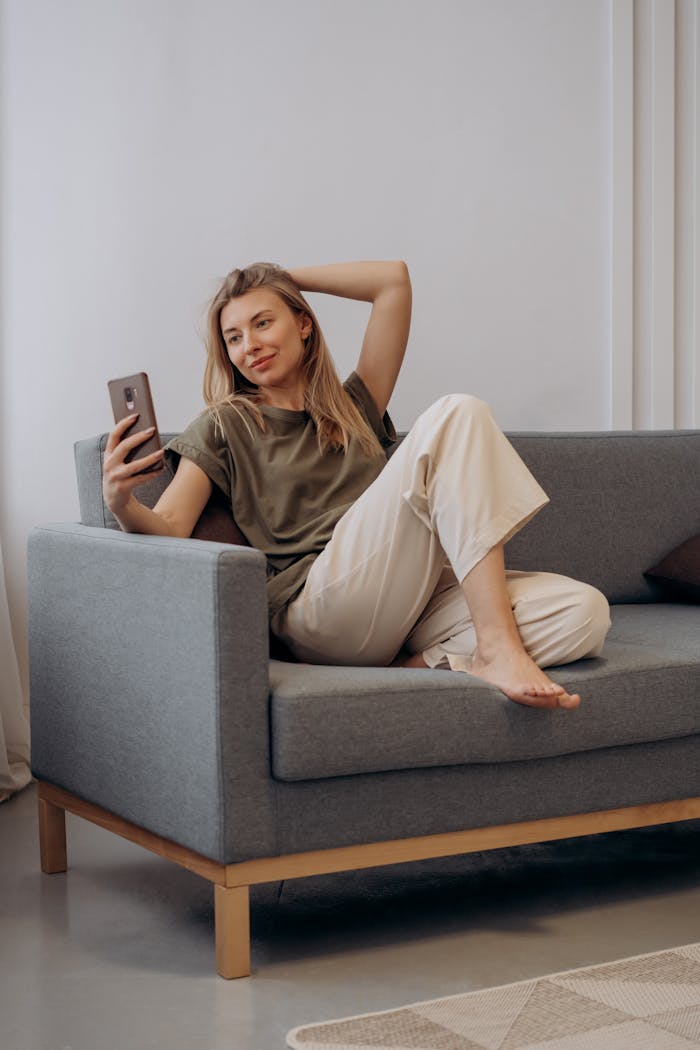 Woman lounging on sofa, taking a selfie with a smartphone, enjoying leisure indoors.