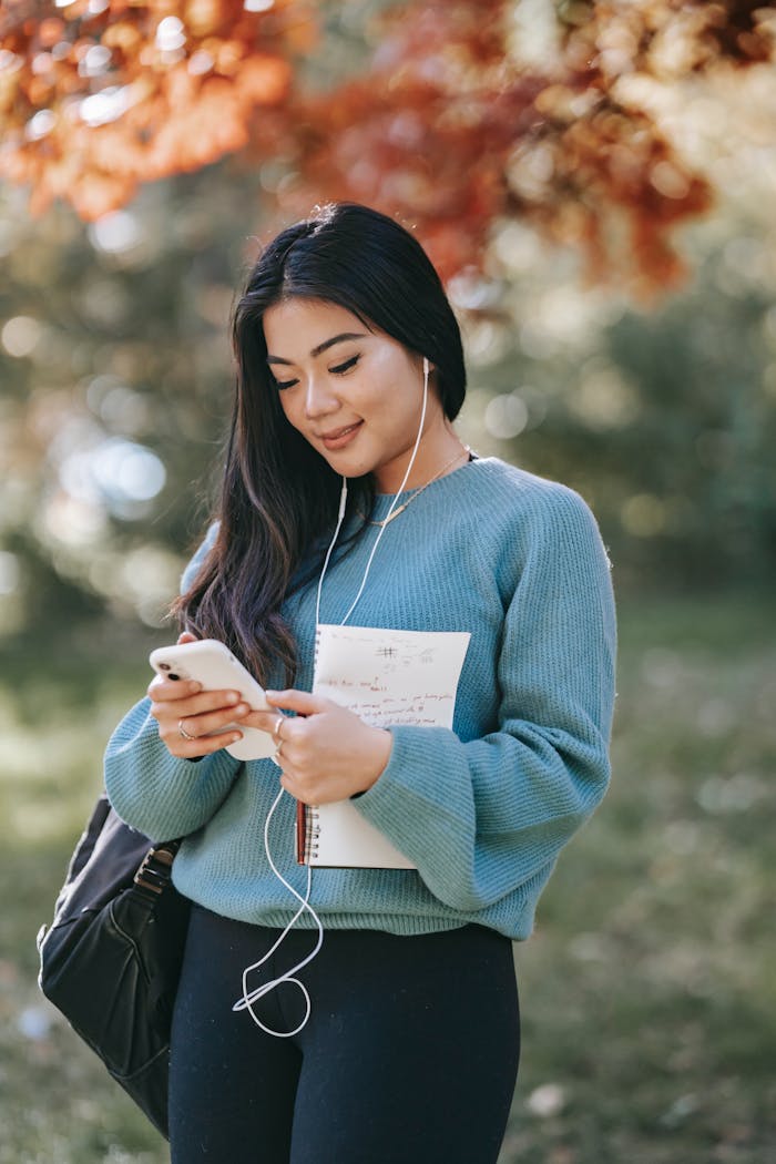 Smiling woman with earbuds using smartphone and notebook in a sunny park.